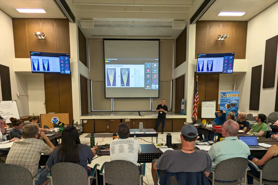 Justin Kay stands in front of a projected presentation in a conference room, addressing attendees.