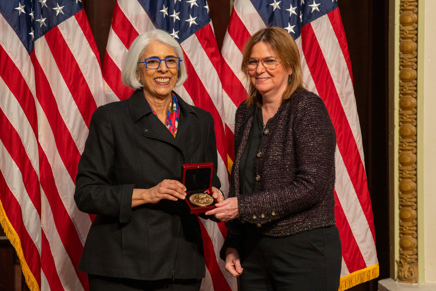 Arati Prabhakar and Angela Belcher pose together with a medal, in front of a U.S. flag