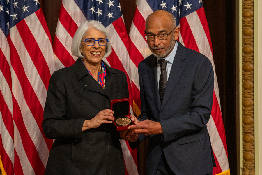 Arati Prabhakar and Emery Brown pose together with a medal, in front of a U.S. flag