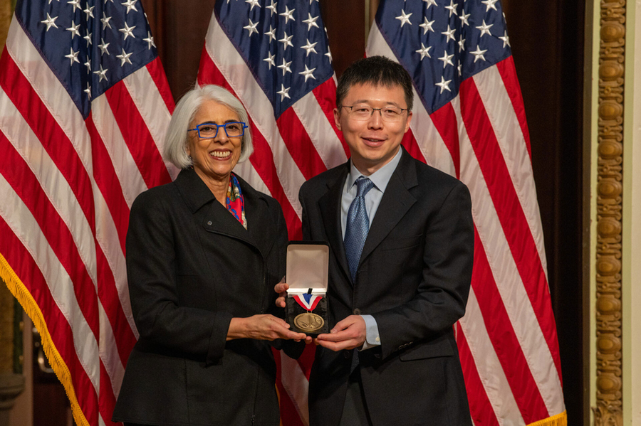 Arati Prabhakar and Feng Zhang pose together with a medal, in front of a U.S. flag