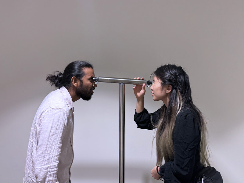  Two people stand facing each other, looking through a pair of eyepieces on opposite ends of a tube-like device. Both are positioned at eye level with the device.