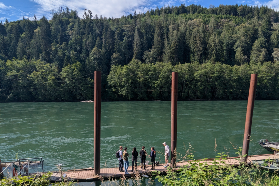 Six people in conversation stand on a dock next to a river.