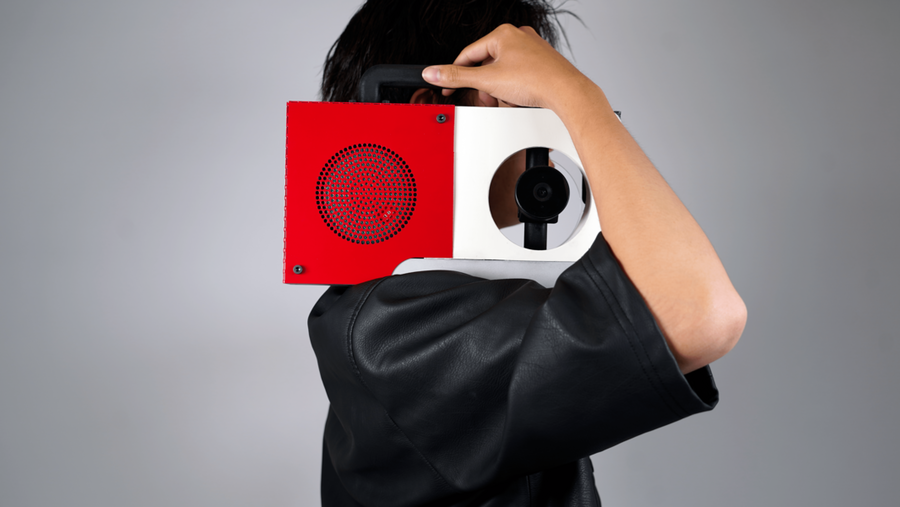 A person whose face is obscured holds on their shoulder a red and white boombox with one speaker replaced with a camera lens.