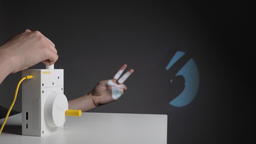 A person operates a white projector device with a crank, labeled "narratron," while their hand creates a shadow of bunny ears projected on a wall.