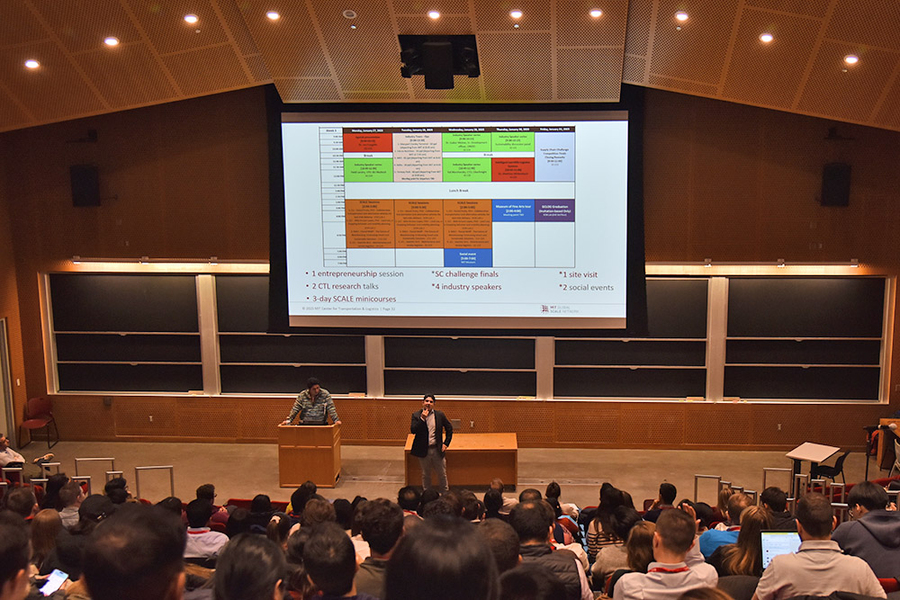 Chris Mejía speaks beneath a scheduling grid projected on a screen in a large lecture hall