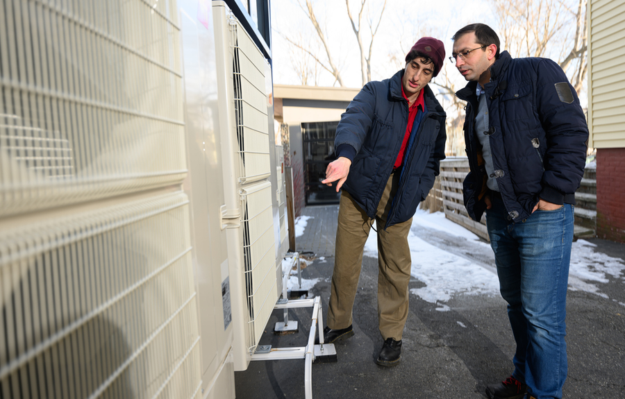 Morgan Santoni-Colvin and Rahman Khorramfar examine a heat pump installed on a home. Snow is on the ground behind them.