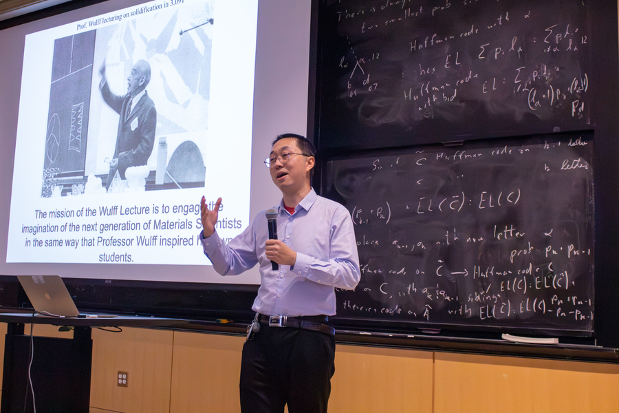 Juejun Hu, standing in front of a blackboard and a video screen at an auditorium at MIT, introduces Vanessa Chan.