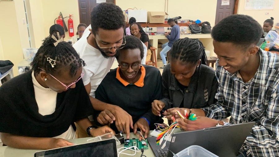 Five students gather around a table with an open laptop and parts needed to build a medical device