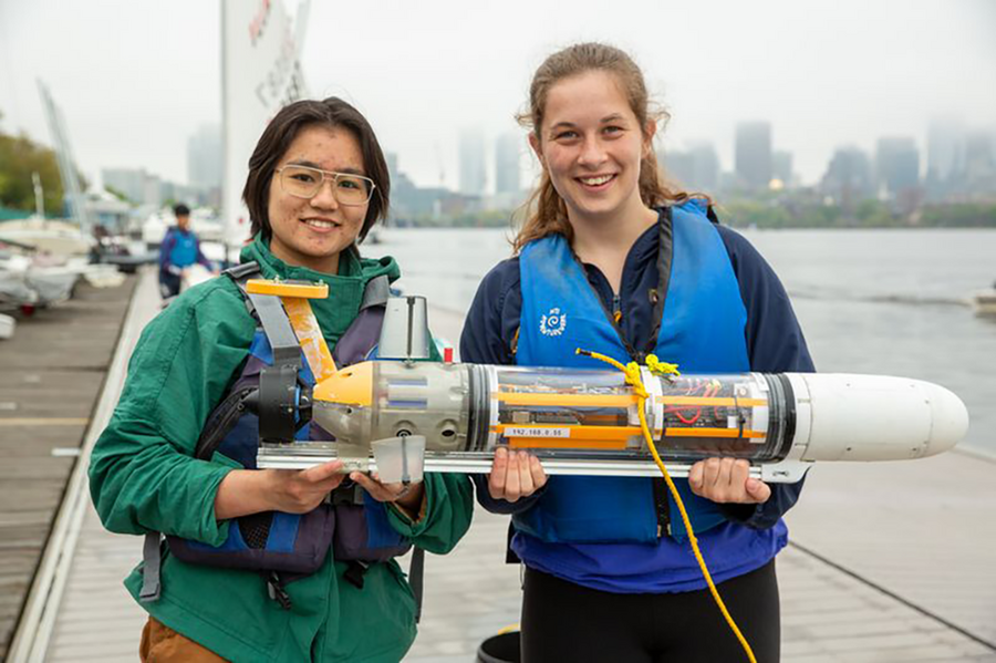 A pair of MIT students stand on a dock next to the Charles River holding a tube-like underwater vehicle
