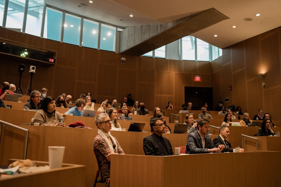 Audience members fill a lecture hall and appear to listen intently to the panel discussions.