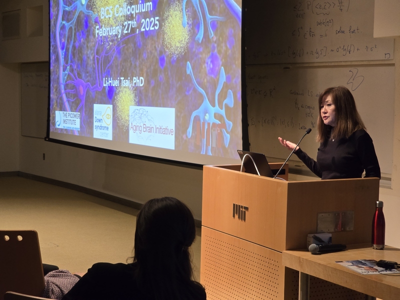Li-Huei Tsai gestures as she speaks at a wooden lectern with an MIT logo in Singleton Auditorium in MIT's Building 46