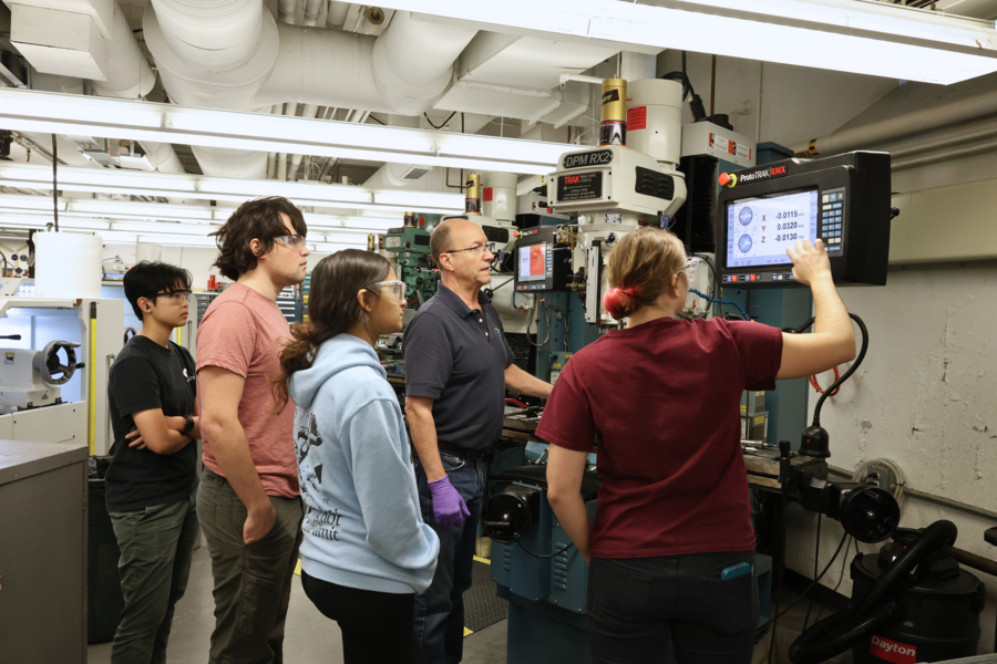 Five students wearing safety glasses in a workshop, listen to an instructor explaining a machine's digital interface.