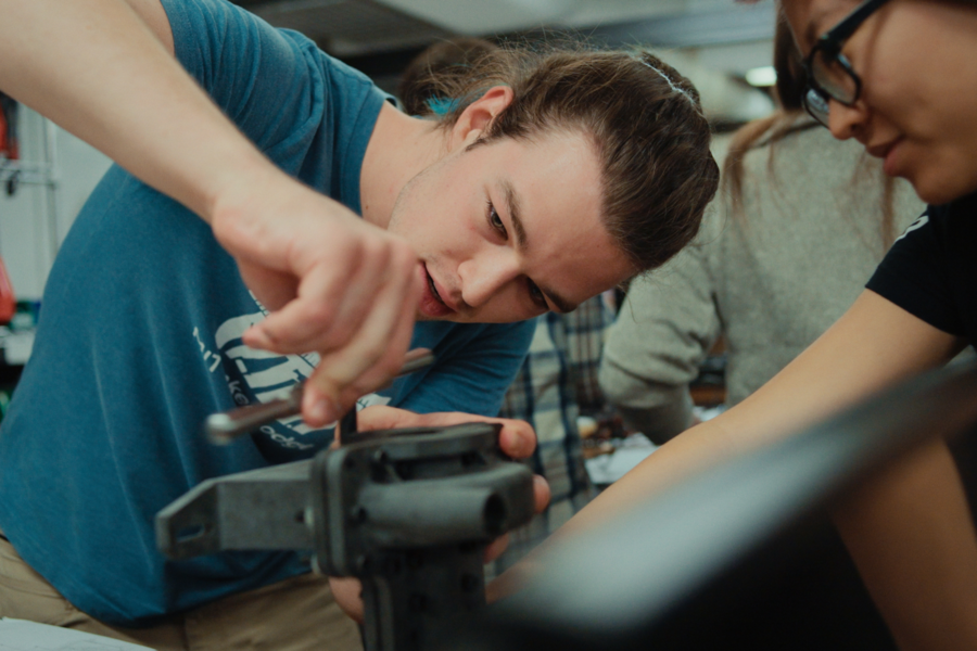 A student works with tools on a mechanical object, surrounded by peers.
