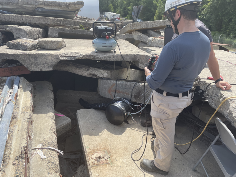 Chad Council stands atop a concrete rubble pile with a controller that controls a robot near a hole in the rubble.