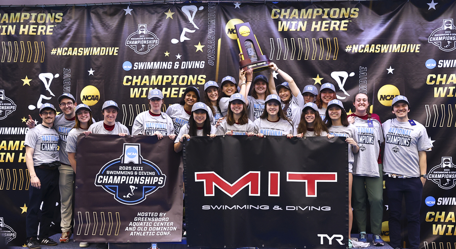 About 20 MIT student-athletes and coaches pose at a podium, hoisting their championship trophy overhead