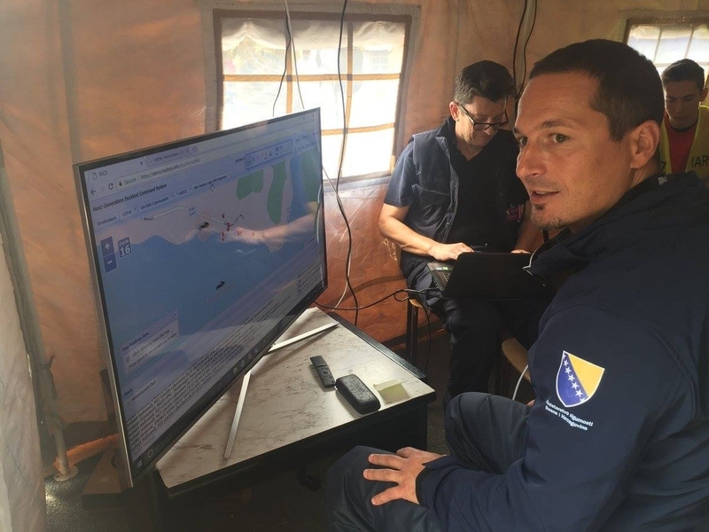 Zeljko Sinkovic sits inside a tent with two others, with a large monitor on a table next to him