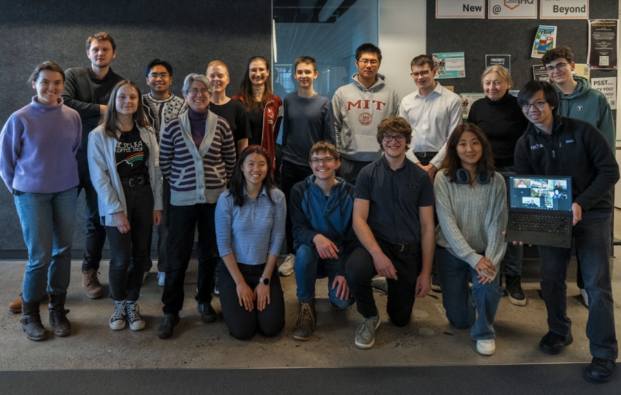 17 people pose in two rows for a group photo. One holds up a laptop screen to include half-a-dozen remote participants.
