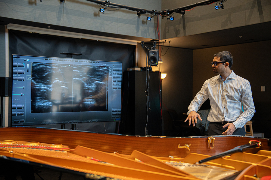 Praneeth Namburi, standing next to a piano, looks at a large screen with two biomechanics images.