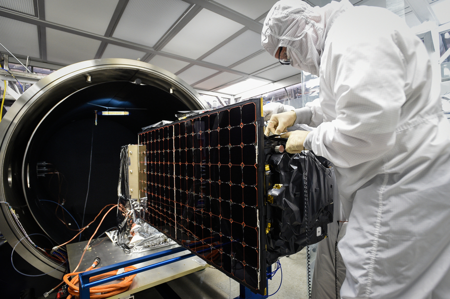 A researcher in a clean suit makes adjustments to a satellite before loading it into an environmental test chamber.