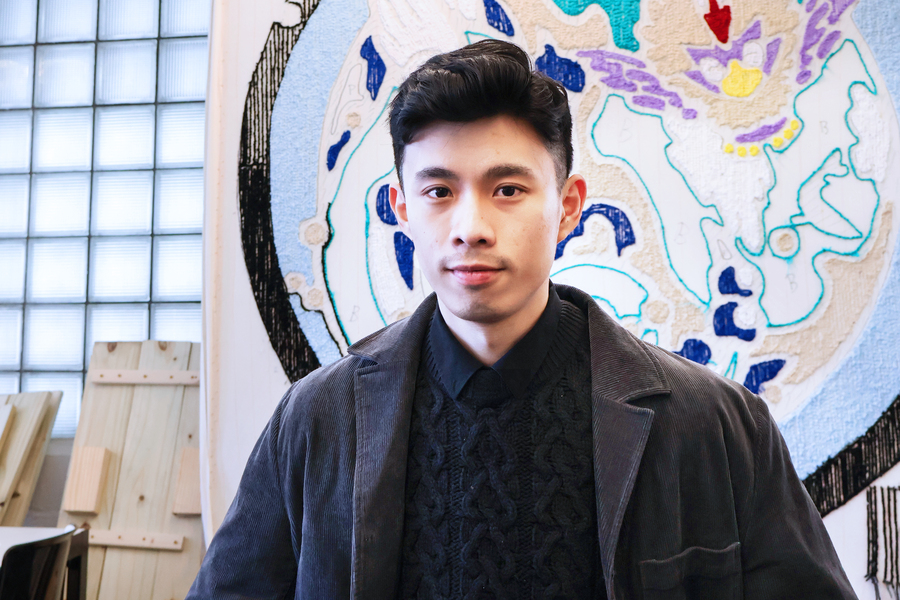 A young man with short black hair wearing a dark cable-knit sweater, black collared shirt, and corduroy jacket stands in front of a large abstract textile artwork featuring bold shapes in blue, white, and purple.