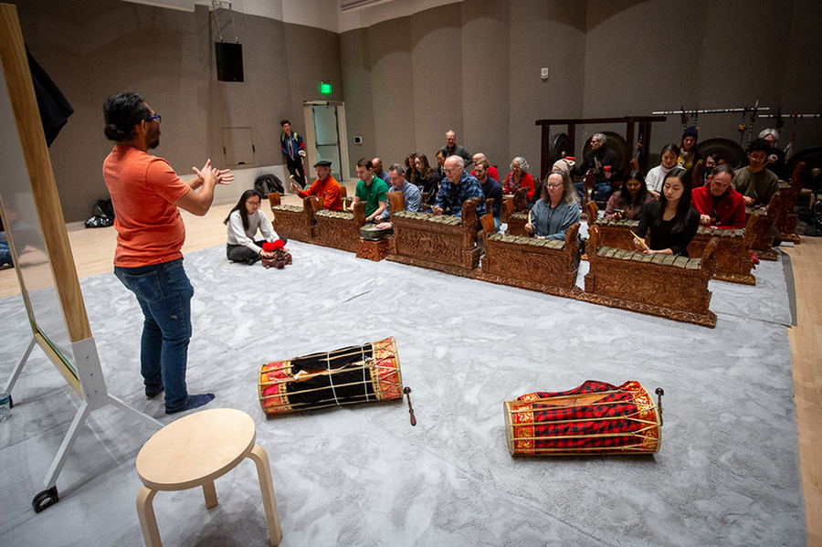 A couple dozen people sit before gamelan instruments in a music room, while a standing individual provides instruction