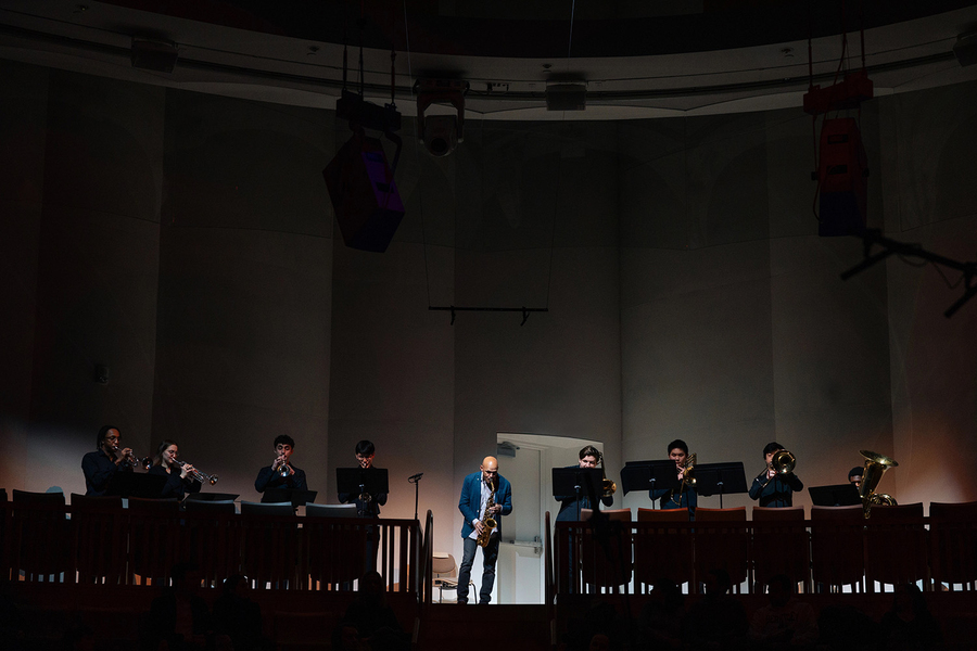 Miguel Zenón plays saxophone with 8 other members of a jazz band in a darkened performance space