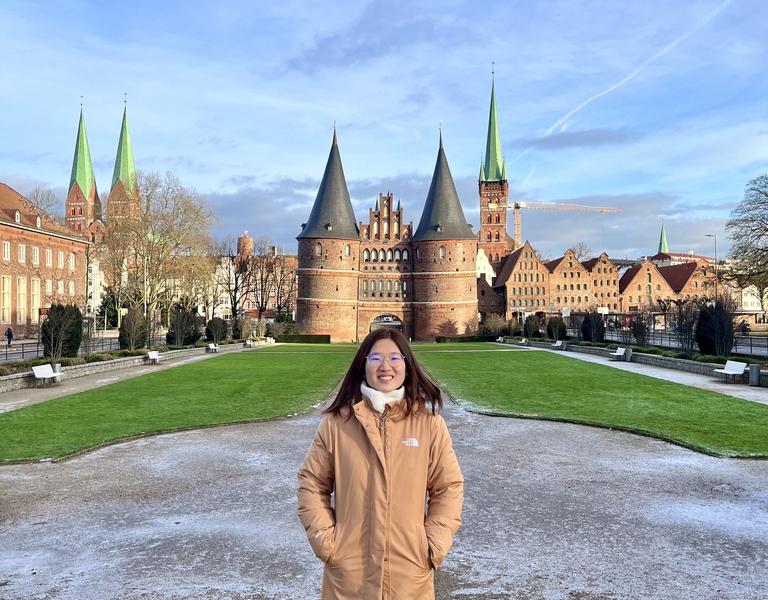 Crystal Liang, wearing a winter jacket, stands in front of a German castle with round towers and high, pointed spires