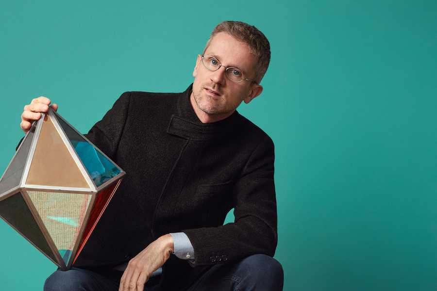 Carlo Ratti poses in a teal room with a geometric figure balanced on his knee