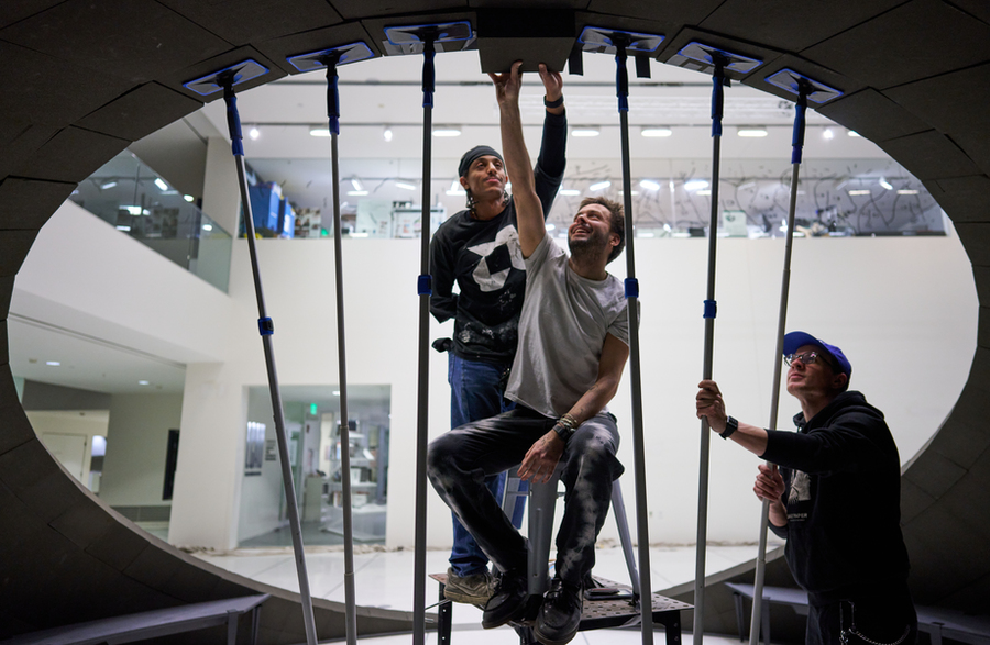 Three people work together to install or adjust a tile on the interior of a large curved structure. One person sits on a ladder, reaching upward, while the others assist using long support poles.