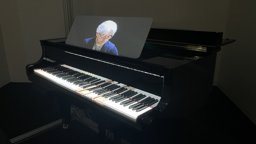A spotlit piano in a darkened room with some keys depressed, and a screen showing a man looking down at the keyboard while playing