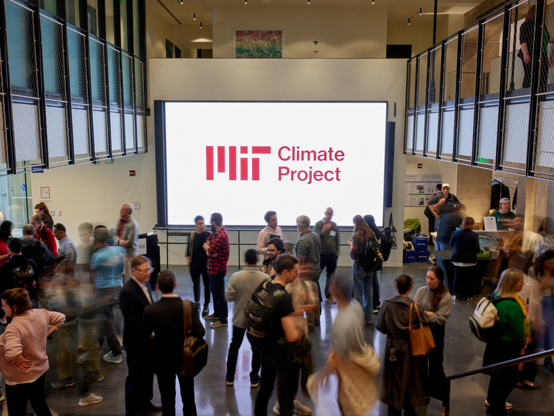 A crowd of people mill about an atrium in front of a large screen with the MIT Climate Project logo.