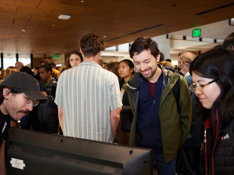 A bearded man smiles in the center of the photo as two people to either side show him something on a computer monitor.