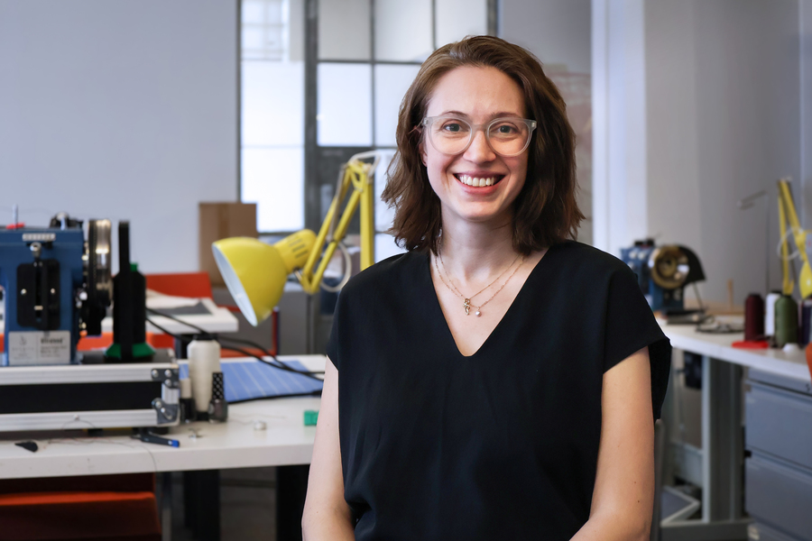 Caitlin Morris smiles while seated in a makerspace at MIT, with tools, a yellow desk lamp, and design equipment visible in the background.