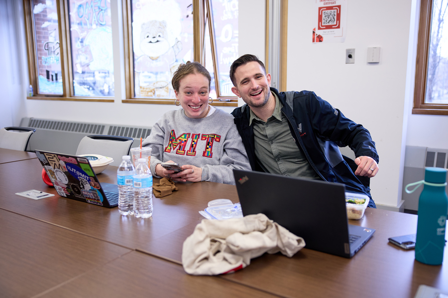 With his arm around another Sloan student, David Brown smiles at the camera. They appear to be eating lunch at their computers.