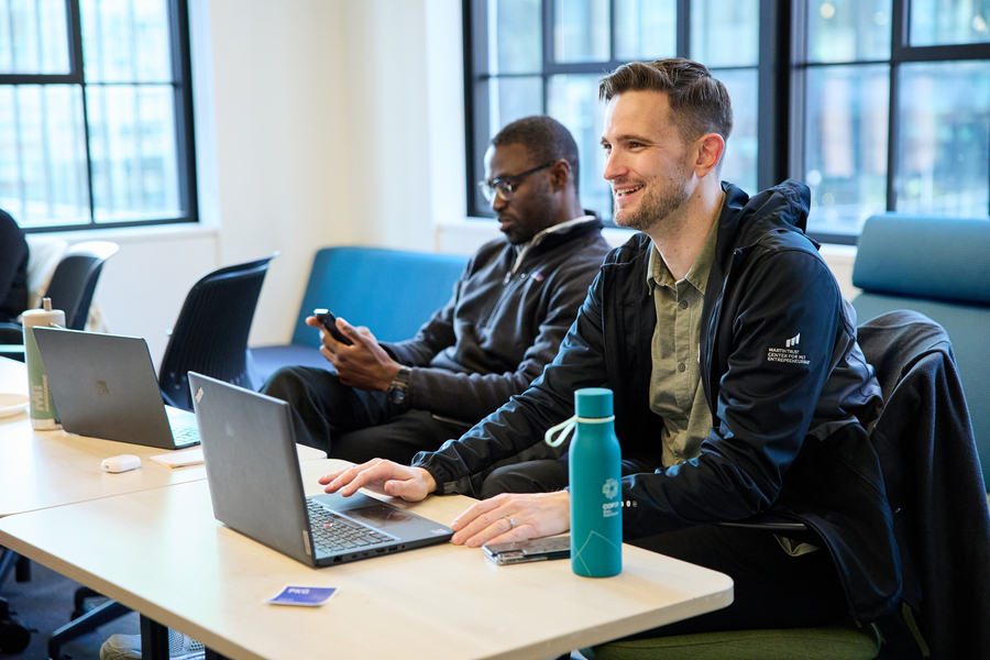 David Brown works at his laptop on a long table where another person is also seated, absorbed in something on his phone
