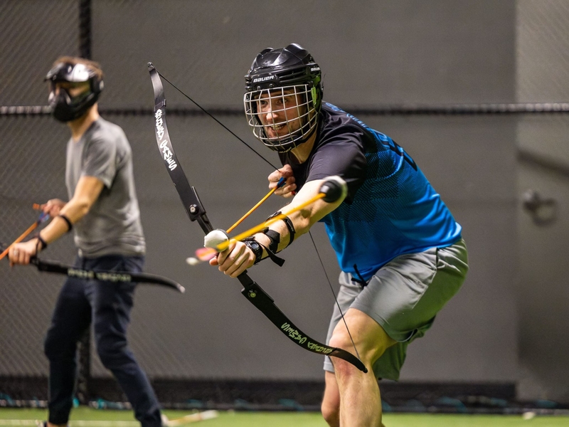 David Brown wears athletic clothing and stands in an indoor athletic facility. He wears a helmet and holds a bow and an arrow with a soft, round ball on the end. He draws back the arrow as if to fire.