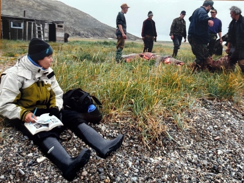 Usha Lee McFarling, dressed in winter clothing, sits by the water with a book and a pencil. To her left, six men stand over an animal carcass.