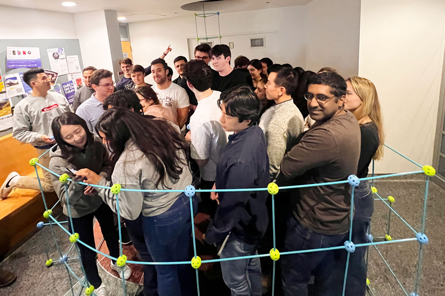 About 20 people huddle together inside a colorful plastic structure made of rods and balls.