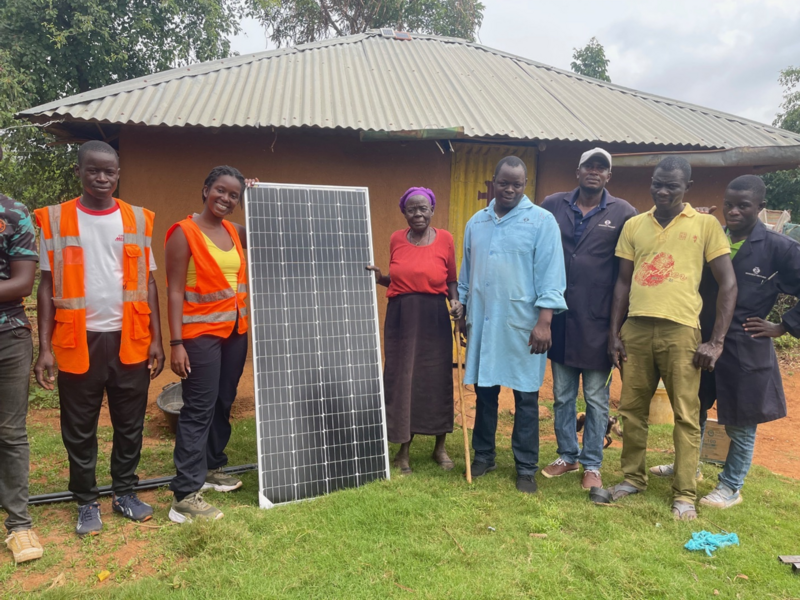 8 people pose standing in front of a small building and showing a solar panel. One person is mostly outside of the frame.