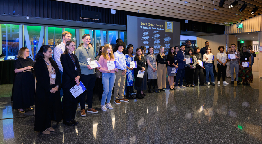 More than two dozen smiling students stand together at the front of a room. Many are holding up certificates.
