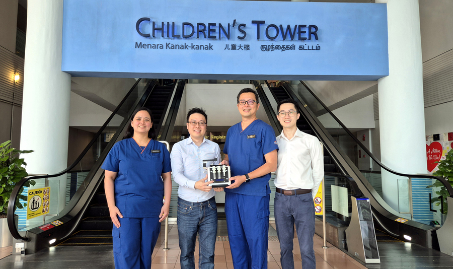 Four people pose in front of two escalators and a sign that says "Children's Tower." The two in the center hold a toaster-sized device.