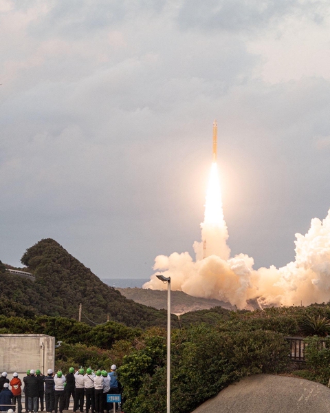 Spectators on a platform watch a rocket launch in the distance over a forest.