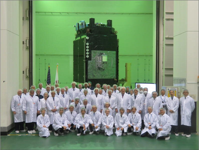 About 50 researchers in white lab coats and hairnets pose in a greenish room with a large satellite in the background.