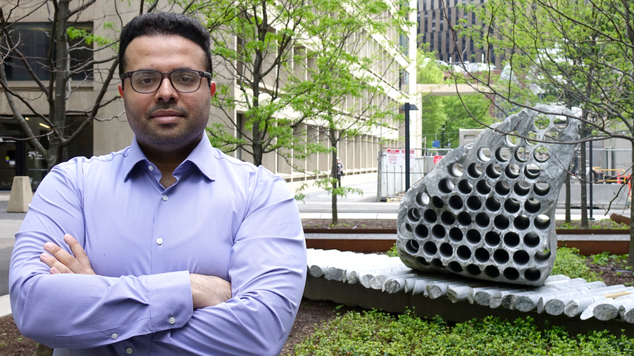 Soroush Mahjoubi stands in front of a concrete building and a concrete-based artwork that looks a bit like a giant honeycomb