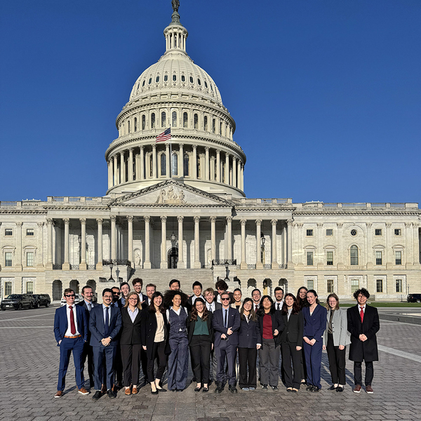 26 people pose together in front of the U.S. Capitol on a sunny day.