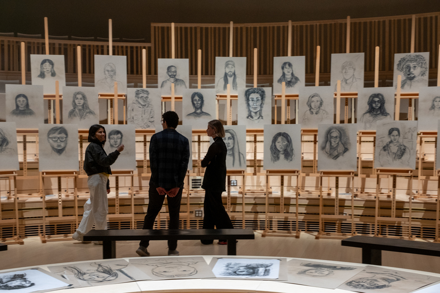 Es Devlin, Perry Naseck, and Sloan Aulgur look at rows of black-and-white portrait drawings on easels. 