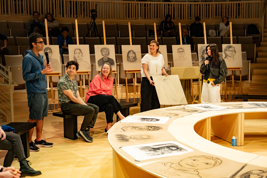 Five people behind a circular table bearing portraits, and in front of eight easels holding more portraits
