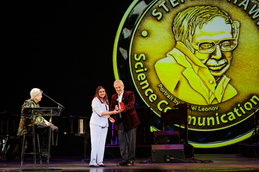 A seated woman and a standing man present an award to a young woman on stage. Behind them is a giant replica of the medal featuring a portrait of Stephen Hawking.