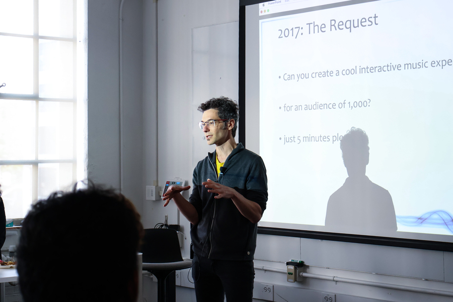 Eran Egozy stands in front of a projected presentation slide, speaking to an unseen audience in a classroom.