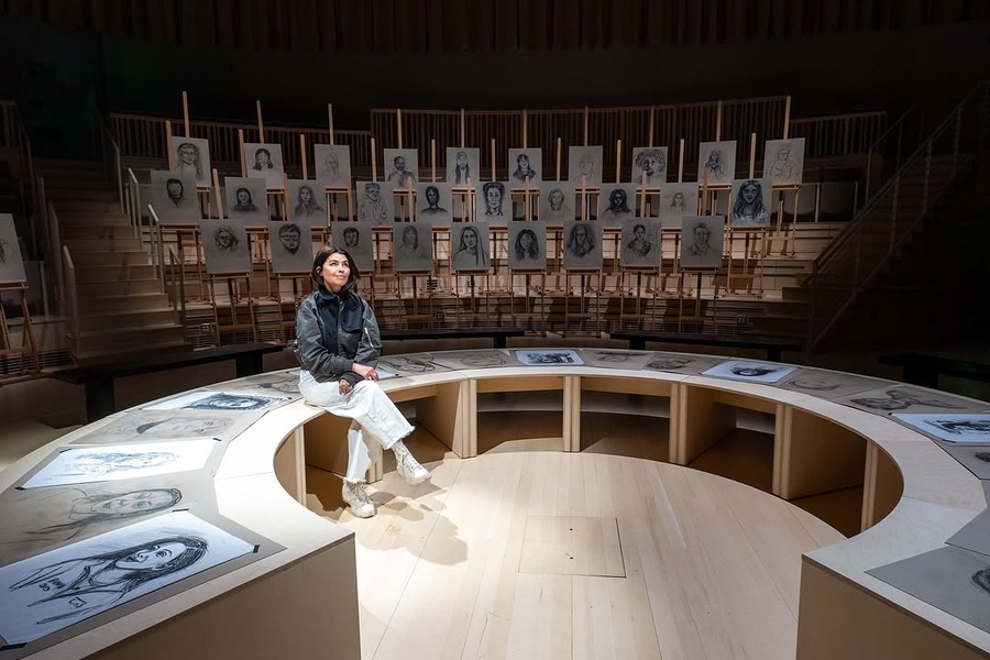 Es Devlin sits on a semicircular bench covered in portrait drawings. 29 more portraits on easels are displayed behind her